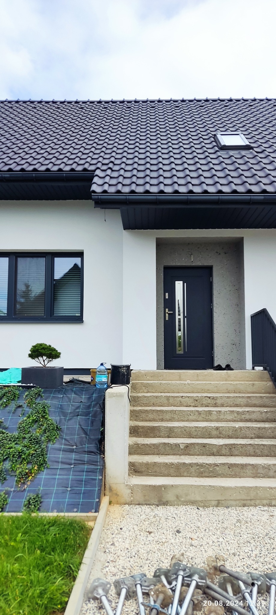 Nowoczesny dom jednorodzinny z białymi ścianami, grafitową stolarką i schodami wejściowymi. Widoczne narzędzia i materiały budowlane.