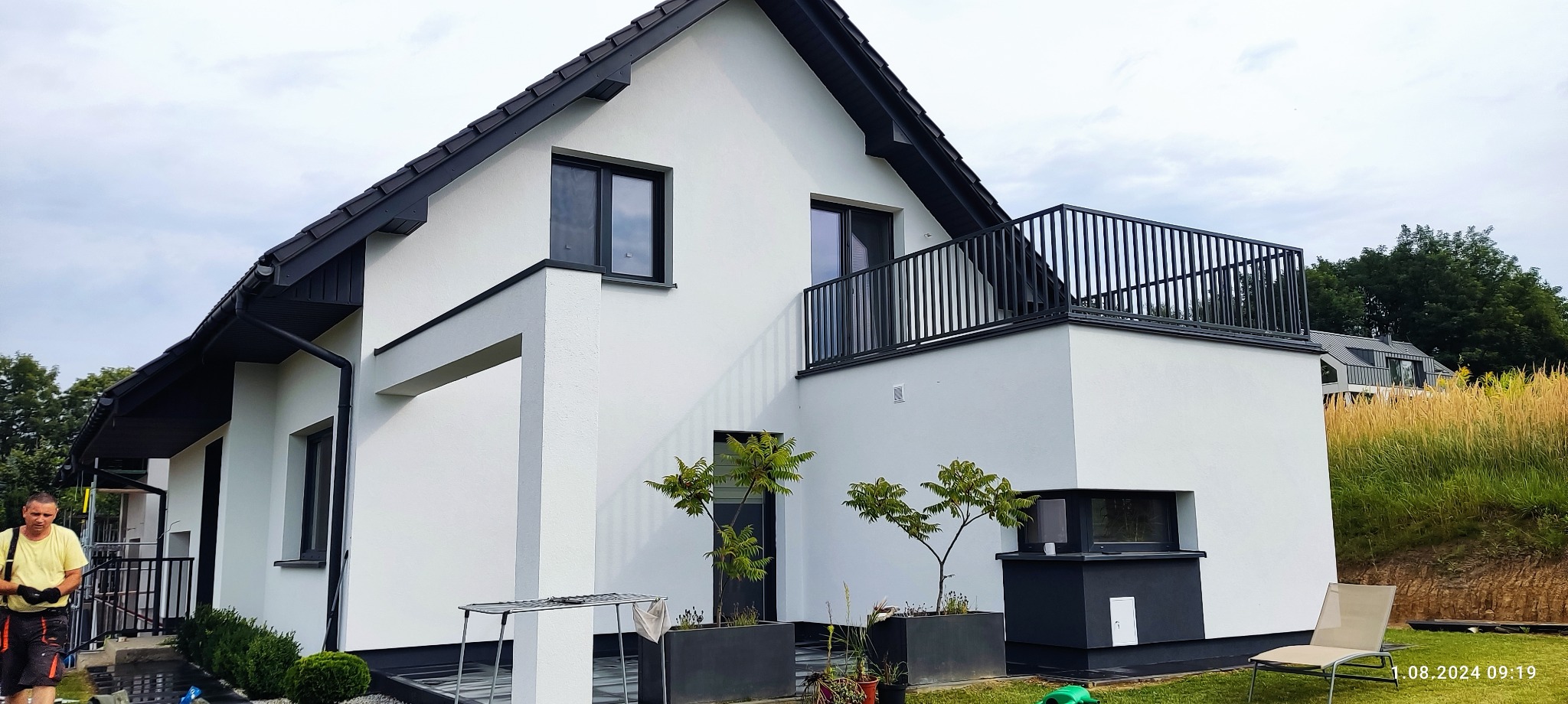 Nowoczesny dom z białą elewacją i czarnym dachem, balkon z metalową balustradą, widok z boku, zieleń w tle, pracownik w tle po lewej.