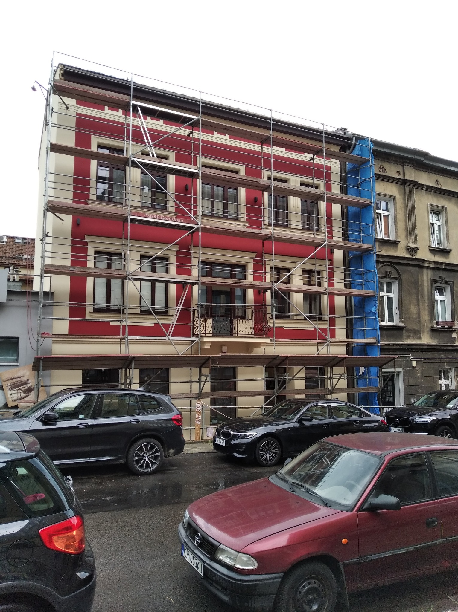 Odnowiona kamienica z rusztowaniem, pomalowana w czerwono-kremowe pasy, sąsiadująca z budynkiem w trakcie renowacji, na mokrej ulicy zaparkowane samochody.