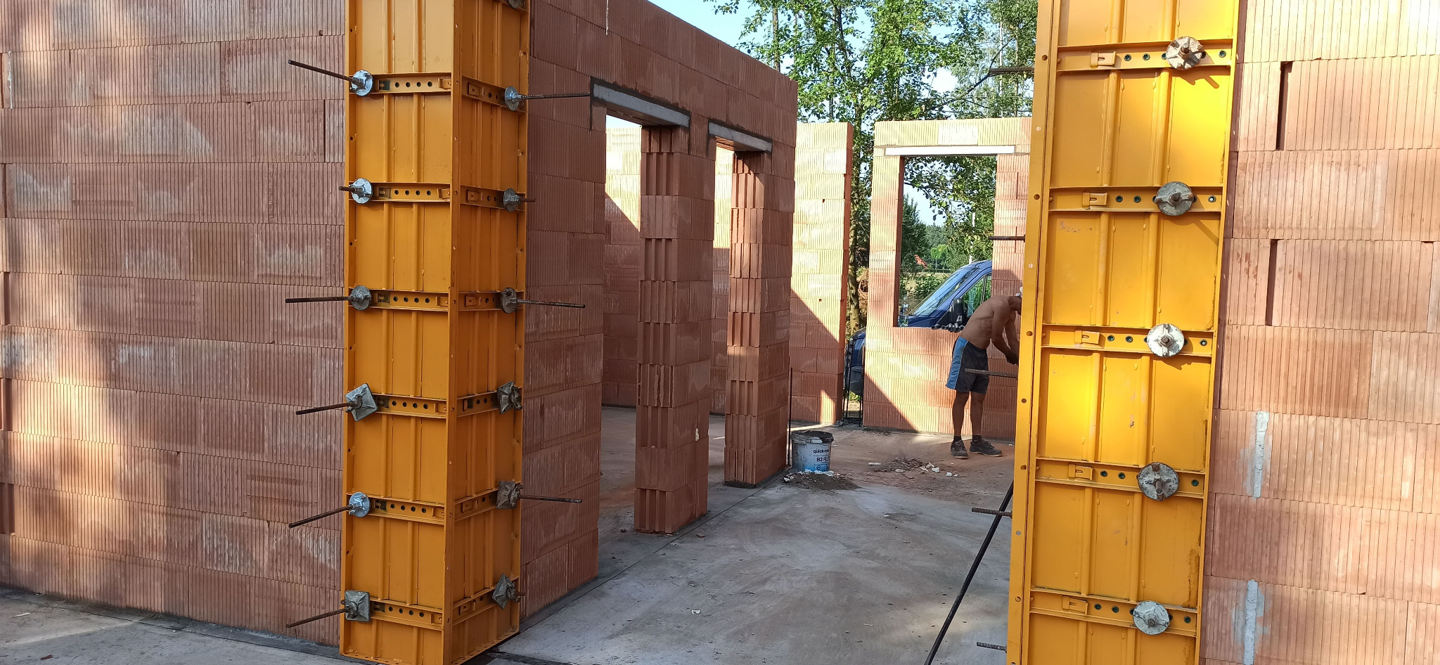 Wznoszenie ścian domu z pustaków ceramicznych, widoczne żółte szalunki, pracownik w tle, wiaderko z zaprawą.