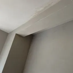 Wnętrze w trakcie prac wykończeniowych: szare, gładkie ściany po nałożeniu gładzi, surowa cegła wokół otworu drzwiowego, widok na okno w tle.