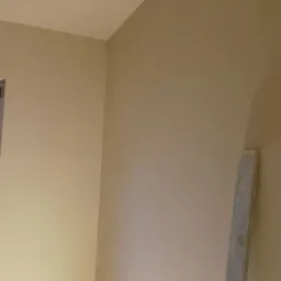 Świeżo otynkowana ściana wewnętrzna pomalowana na beżowo, widoczna poziomica oparta o ścianę w prawym dolnym rogu, oświetlenie sufitowe tworzy cień na górnej części ściany.