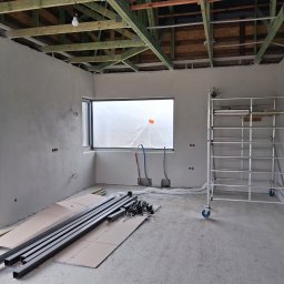 KES-TYNK Sebastian Adamczyk Usługi Tynkarskie - Wnętrze w trakcie prac wykończeniowych: ściany po nałożeniu tynku, widoczne rury instalacyjne, rusztowanie na kółkach i elementy konstrukcyjne na podłodze.