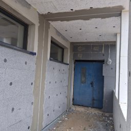 KES-TYNK Sebastian Adamczyk Usługi Tynkarskie - Ocieplenie budynku szarym styropianem grafitowym, widoczne okna i niebieskie drzwi wejściowe. Prace w toku, widoczne narzędzia i resztki materiałów budowlanych.