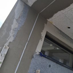 KES-TYNK Sebastian Adamczyk Usługi Tynkarskie - Ocieplenie budynku z widocznym oknem, fragmentem rusztowania i świeżo nałożonym tynkiem. Widoczne detale wykończenia i struktura materiałów.