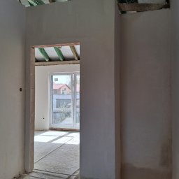 KES-TYNK Sebastian Adamczyk Usługi Tynkarskie - Wnętrze w stanie deweloperskim po tynkowaniu, widok przez otwór drzwiowy na okno z widokiem na budynek. Na podłodze widoczne rurki ogrzewania podłogowego.
