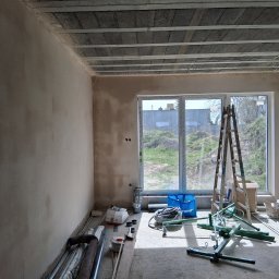 KES-TYNK Sebastian Adamczyk Usługi Tynkarskie - Wnętrze w trakcie prac wykończeniowych: ściana po tynkowaniu, widoczne narzędzia, drabina przy oknie, surowy strop betonowy. Przygotowanie do dalszych prac.