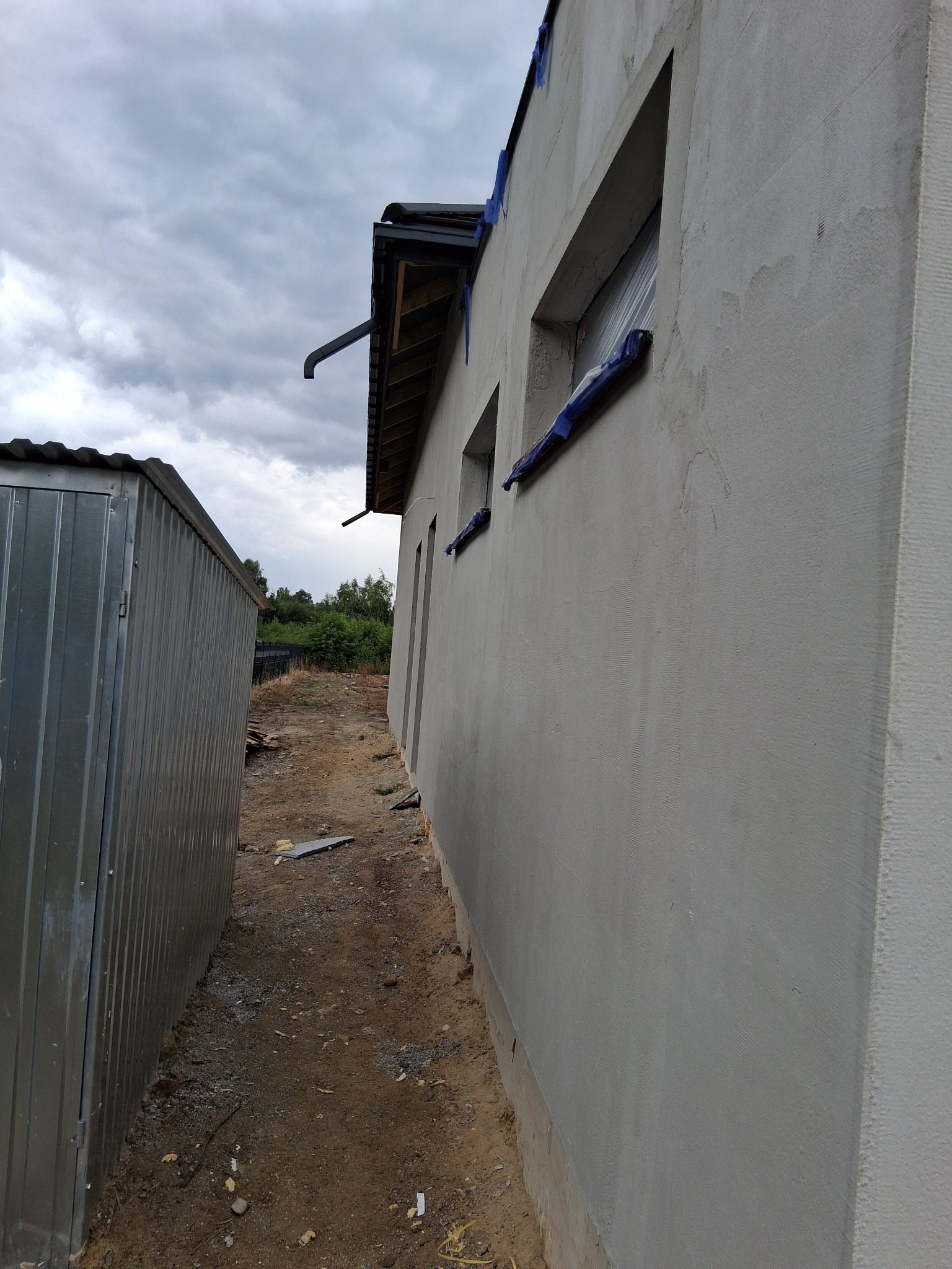 Świeżo otynkowana ściana budynku z zabezpieczonymi oknami, widoczna z boku, obok blaszany garaż i fragment dachu. Pochmurne niebo w tle.