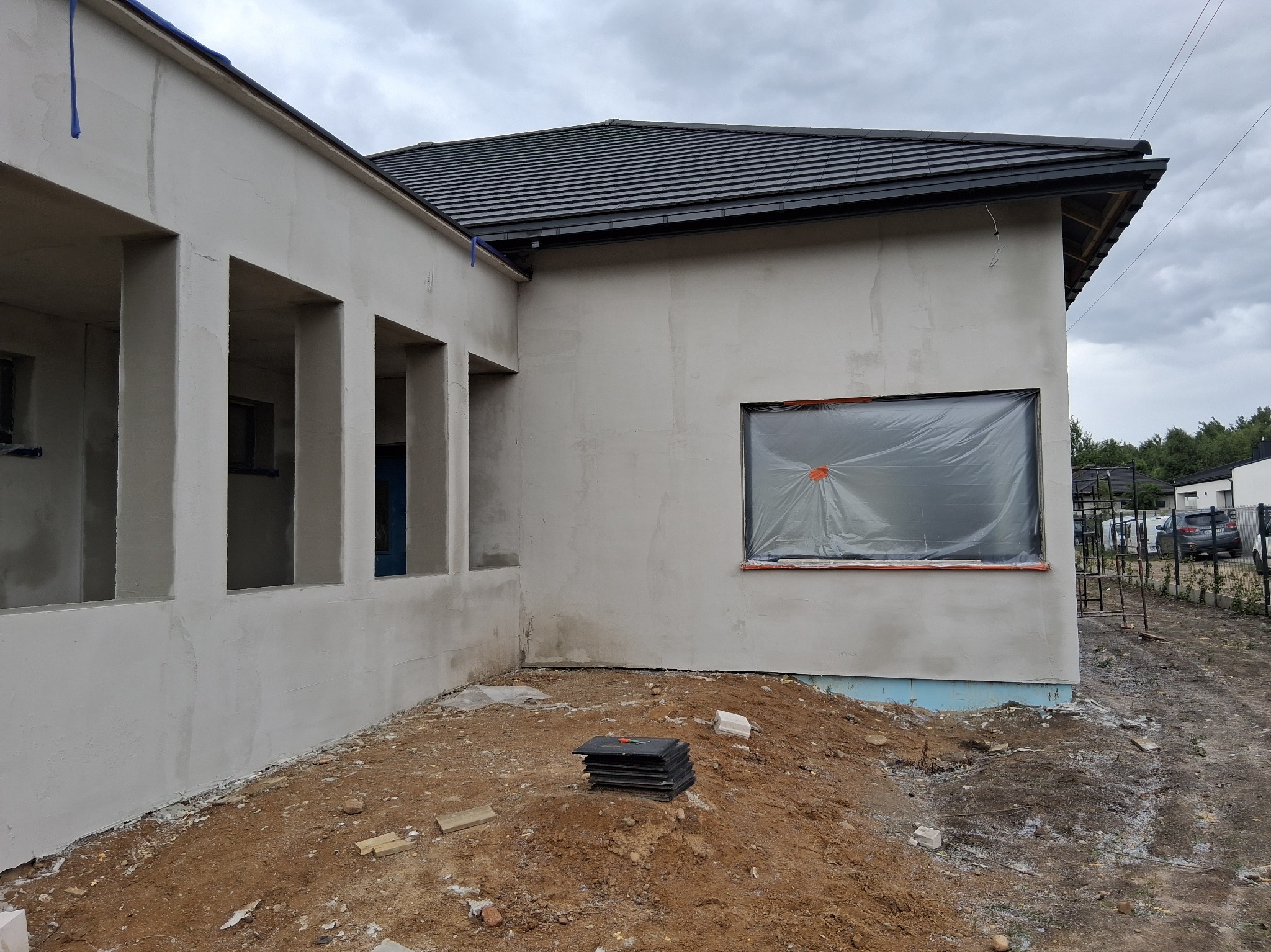 Świeżo otynkowany dom jednorodzinny z czarną dachówką, okno zabezpieczone folią. Widoczny fragment elewacji z kolumnami. Na pierwszym planie ziemia i czarne płyty.