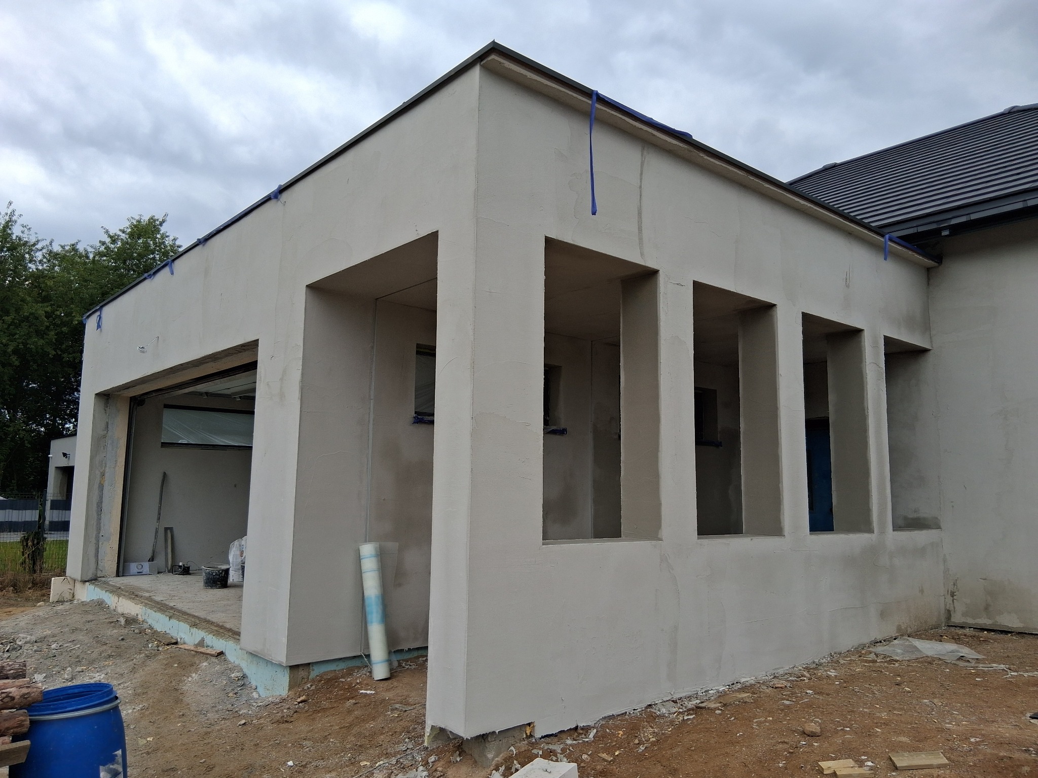 Budynek w trakcie prac tynkarskich: świeżo otynkowana elewacja z widocznymi otworami okiennymi i fragmentem dachu. Niebieska taśma zabezpieczająca na krawędziach.
