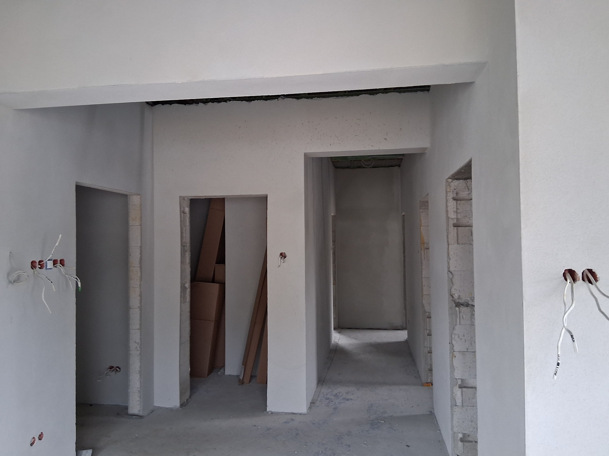 Wnętrze w trakcie prac wykończeniowych: świeżo otynkowane ściany, widoczne instalacje elektryczne, surowy beton na podłodze, przygotowane otwory drzwiowe.