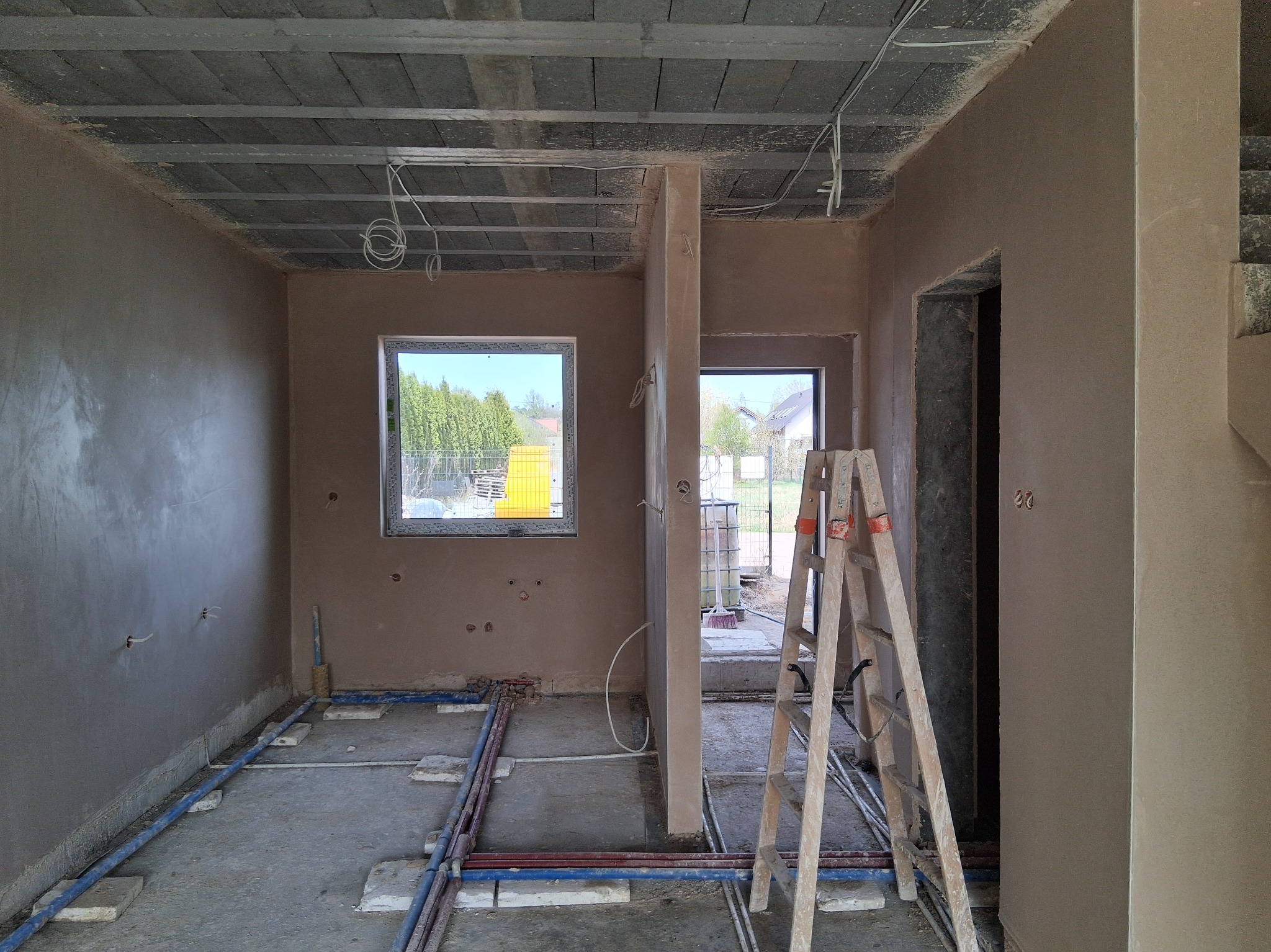 Wnętrze w trakcie prac tynkarskich: ściany pokryte świeżym tynkiem, widoczne instalacje podłogowe i drabina. Surowy strop betonowy z widocznymi przewodami.