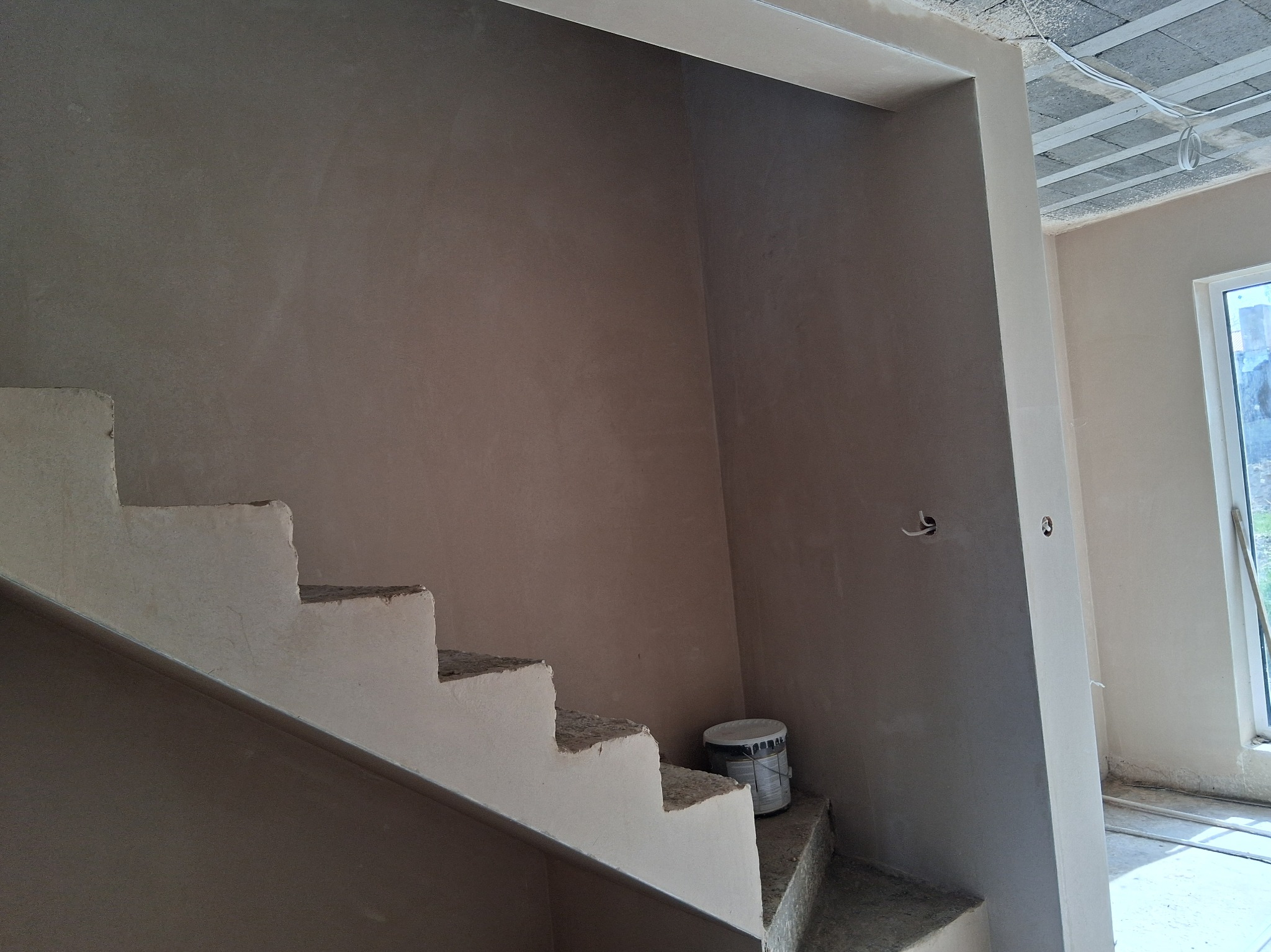 Wnętrze w trakcie prac wykończeniowych: otynkowane ściany, surowy beton schodów, puszka z farbą przy schodach, widoczne okno i instalacje pod sufitem.