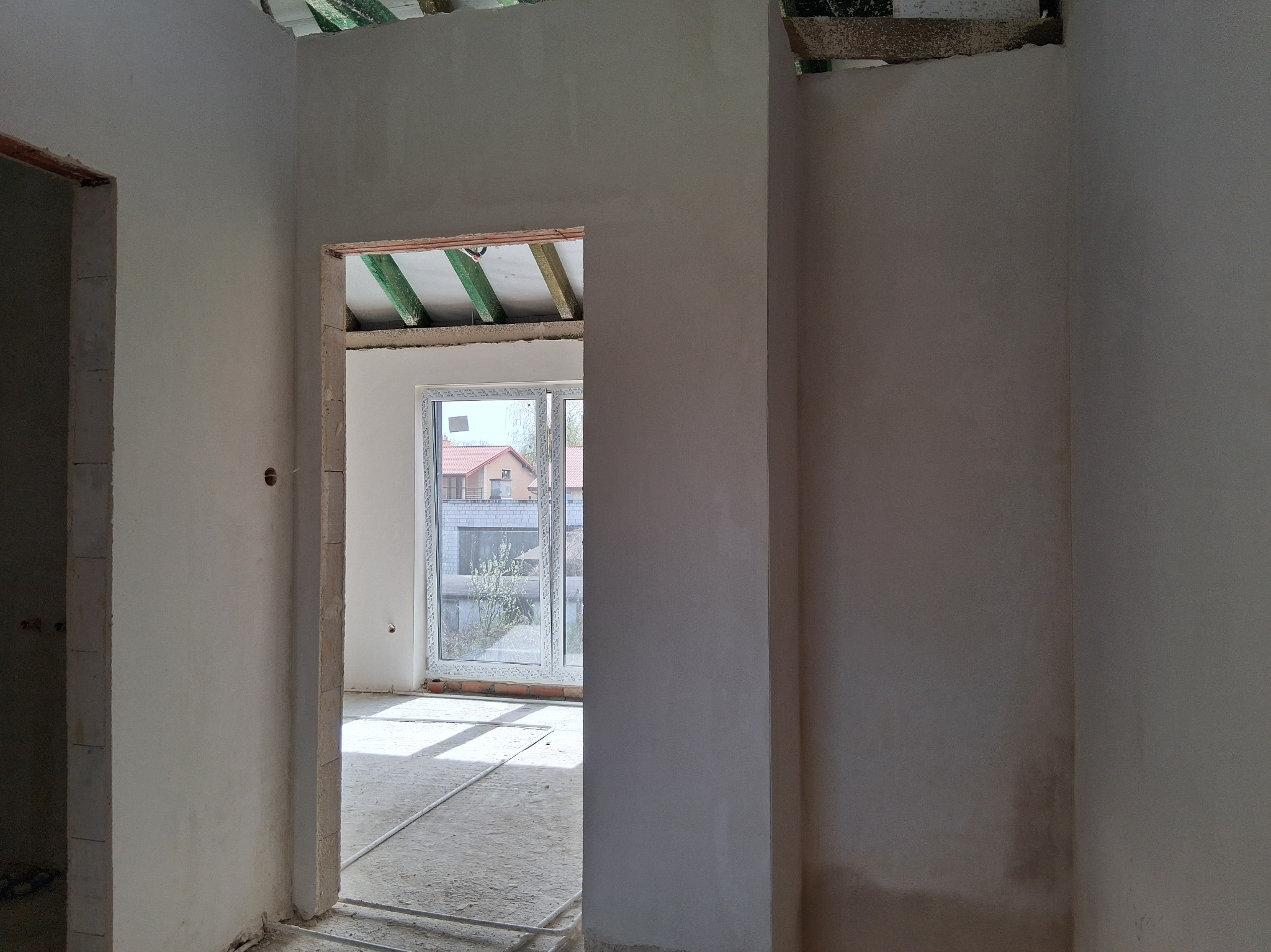 Wnętrze w stanie deweloperskim po tynkowaniu, widok przez otwór drzwiowy na okno z widokiem na budynek. Na podłodze widoczne rurki ogrzewania podłogowego.