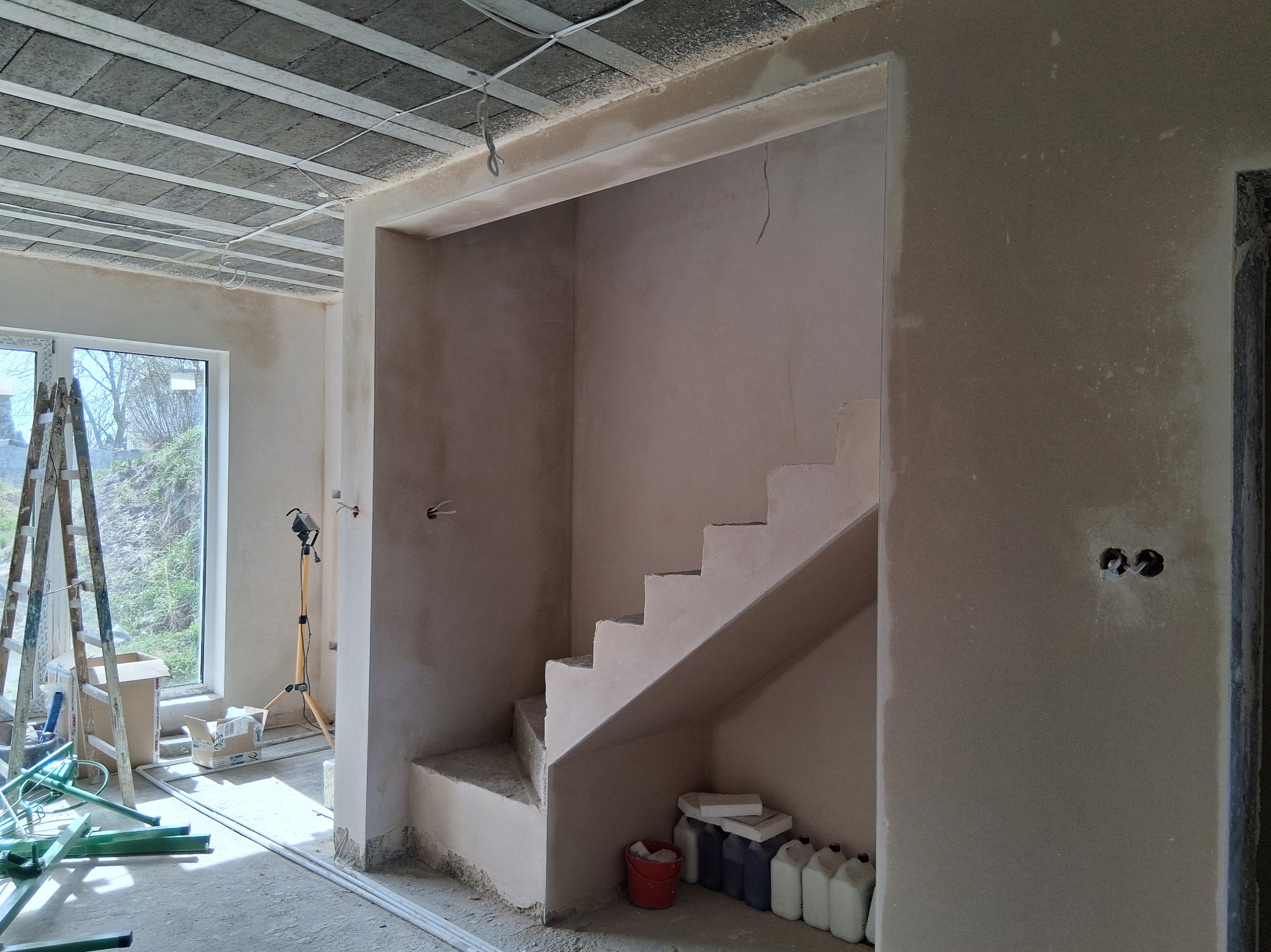 Wnętrze w trakcie prac wykończeniowych: otynkowane ściany, sufit z widoczną konstrukcją, schody w stanie surowym, drabina oparta o okno, widoczne materiały budowlane.