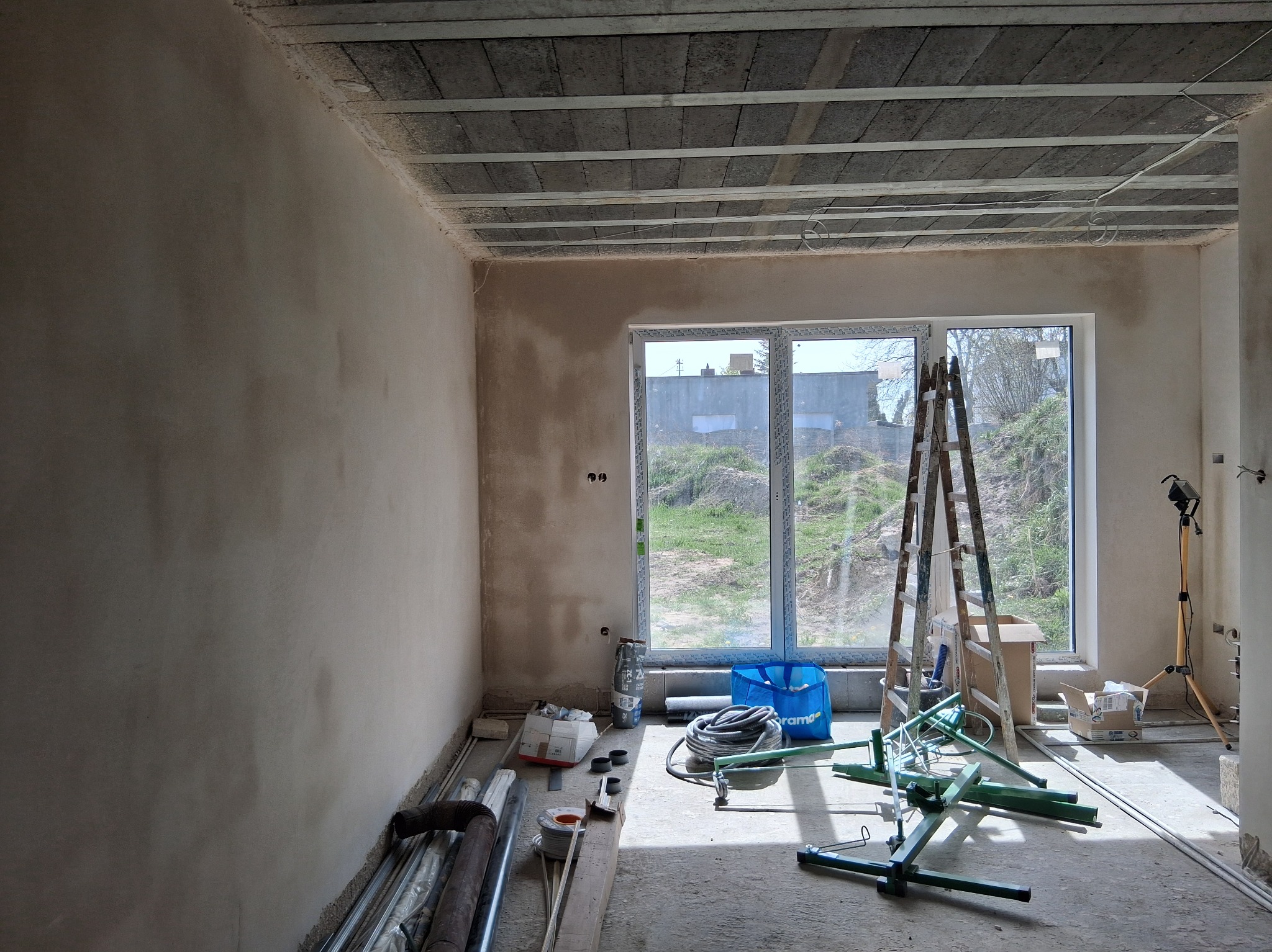 Wnętrze w trakcie prac wykończeniowych: ściana po tynkowaniu, widoczne narzędzia, drabina przy oknie, surowy strop betonowy. Przygotowanie do dalszych prac.