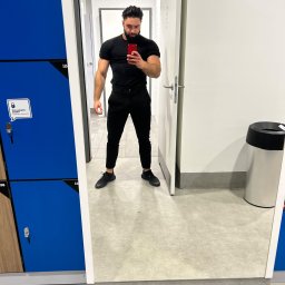 Treningi personalne - Mężczyzna w czarnym ubraniu robi selfie w lustrze w szatni siłowni, widoczne niebieskie szafki i białe drzwi do toalety.