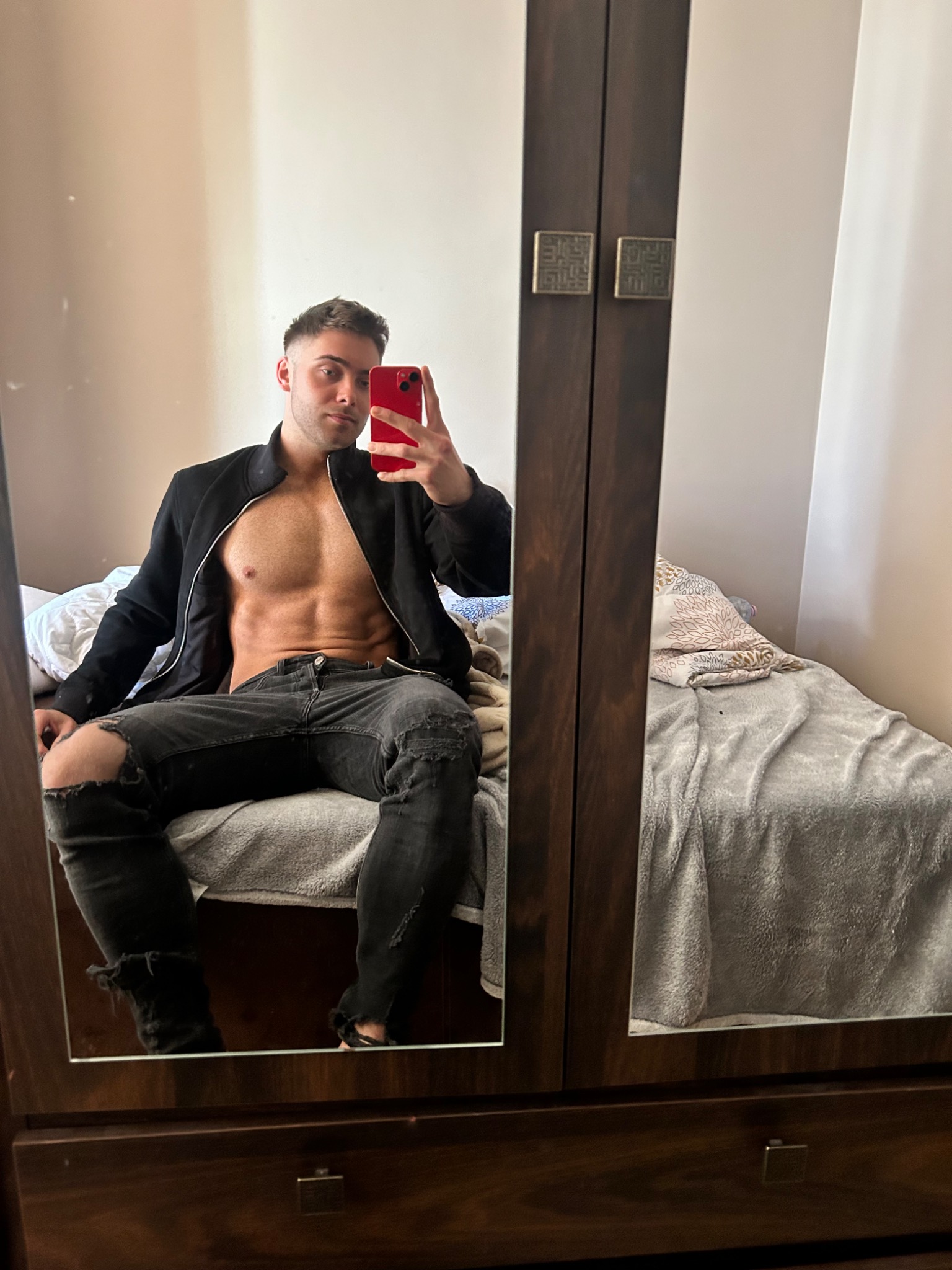 Mężczyzna z widocznymi mięśniami brzucha, w czarnej rozpiętej bluzie i podartych dżinsach, robi selfie w lustrze szafy w sypialni.