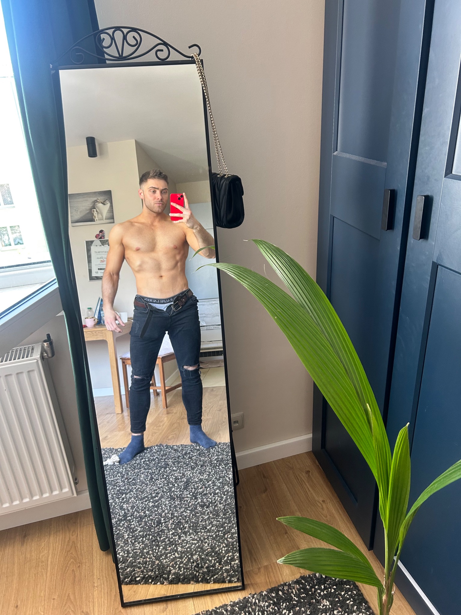 Mężczyzna bez koszulki w jeansach pozuje w lustrze, trzymając czerwony telefon, widoczny fragment wnętrza z rośliną doniczkową.