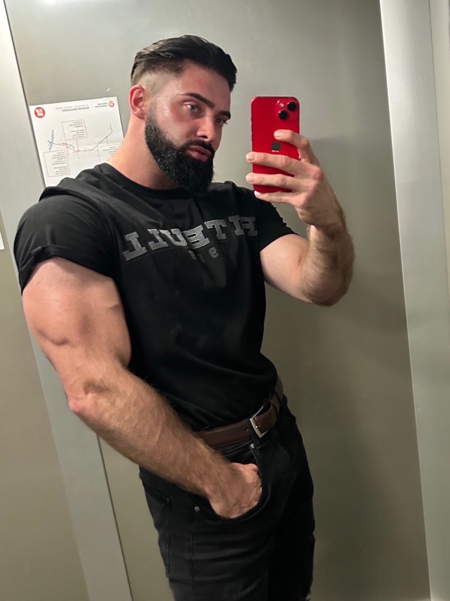 Mężczyzna z brodą robi selfie w lustrze, prezentując bicepsy, ubrany w czarną koszulkę i dżinsy.