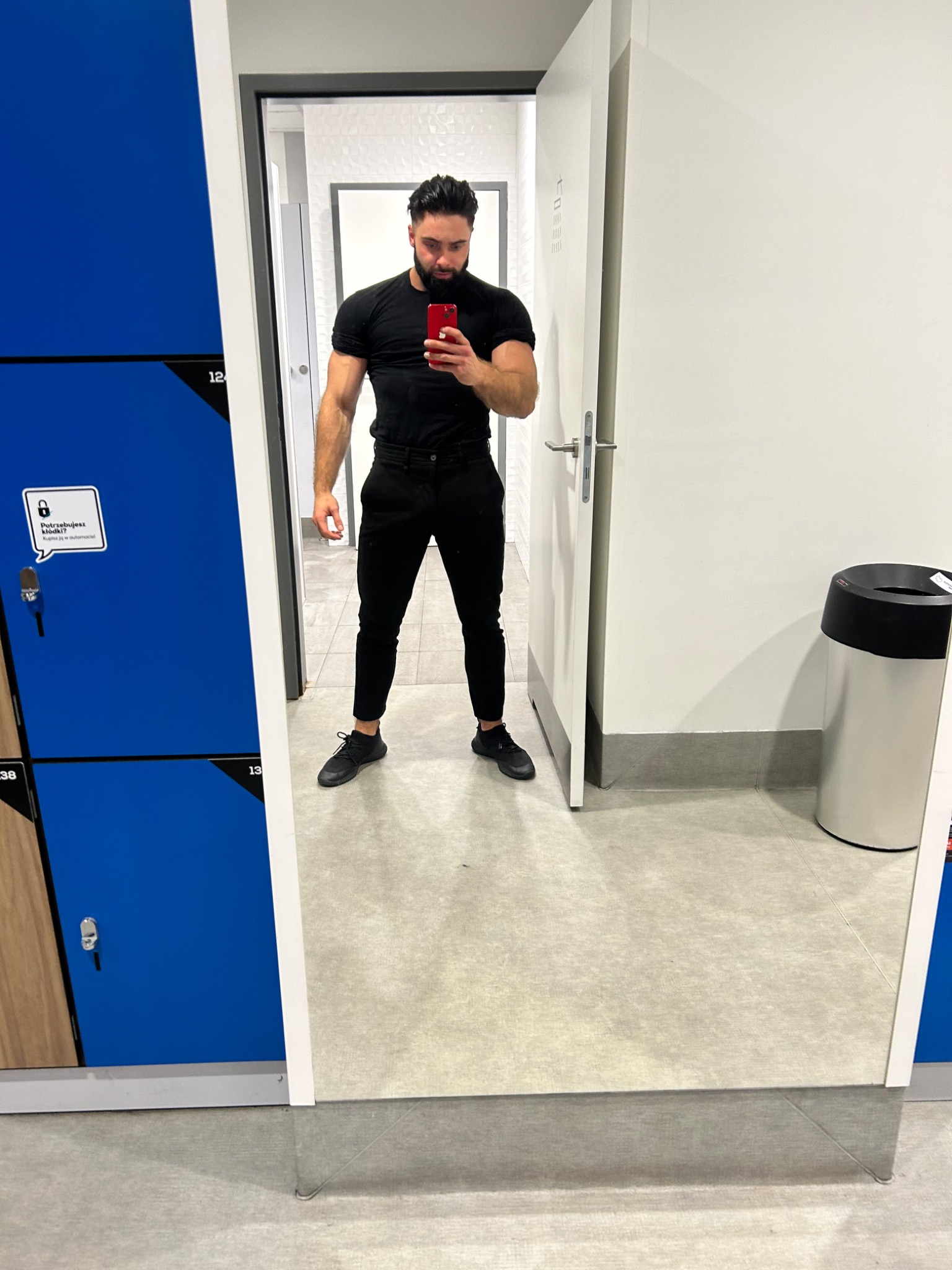 Mężczyzna w czarnym ubraniu robi selfie w lustrze w szatni siłowni, widoczne niebieskie szafki i białe drzwi do toalety.