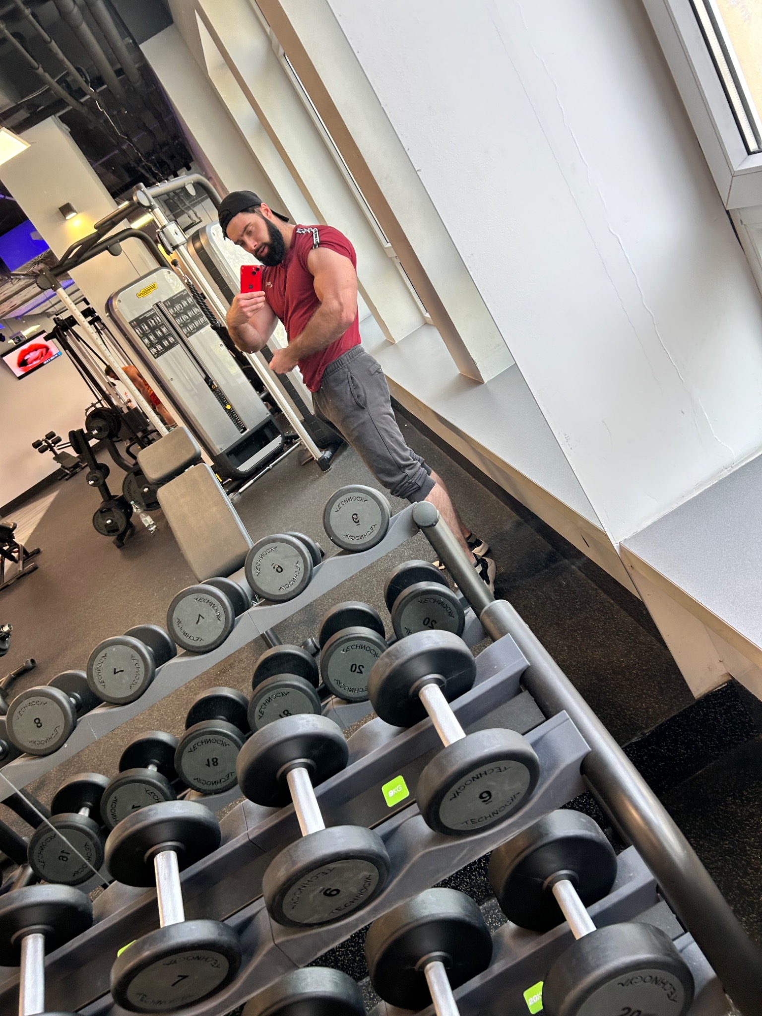 Mężczyzna w czapce i sportowym stroju robi selfie w siłowni, prezentując bicepsy na tle stojaka z hantlami i maszyn do ćwiczeń.