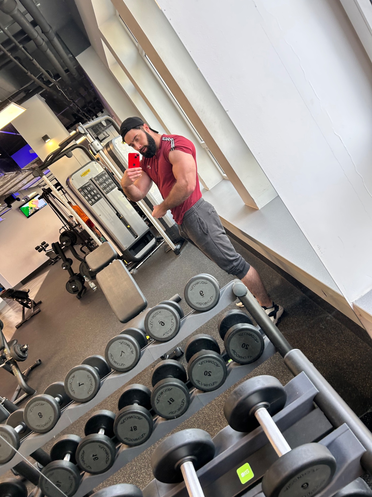 Mężczyzna w sportowym stroju robi selfie w siłowni na tle stojaków z hantlami i maszyn do ćwiczeń.