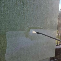 SolarCare BORUSIAK GRZEGORZ - Strumień wody pod ciśnieniem z dyszy lancy czyści fragment zielonkawej, chropowatej elewacji budynku, widoczny kontrast czystej i zabrudzonej powierzchni.