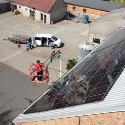 SolarCare BORUSIAK GRZEGORZ - Pracownik na podnośniku koszowym czyści panele słoneczne na metalowym dachu budynku gospodarczego. Na dole widoczny biały samochód dostawczy i sprzęt do czyszczenia.
