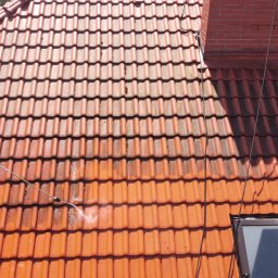 SolarCare BORUSIAK GRZEGORZ - Częściowo wyczyszczony dach z czerwonej dachówki widziany z góry, pracownik na podnośniku koszowym czyści dach myjką ciśnieniową, widoczny komin i okno dachowe.