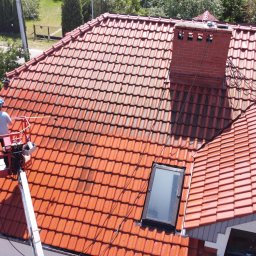 SolarCare BORUSIAK GRZEGORZ - Pracownik na podnośniku koszowym czyści dach z czerwonej dachówki, widoczny fragment dachu przed i po czyszczeniu, ceglany komin.