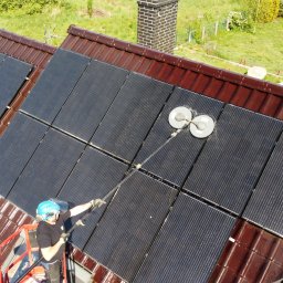 SolarCare BORUSIAK GRZEGORZ - Pracownik w kasku i na podnośniku koszowym czyści panele słoneczne na brązowym dachu za pomocą specjalistycznej szczotki obrotowej, widok z góry.