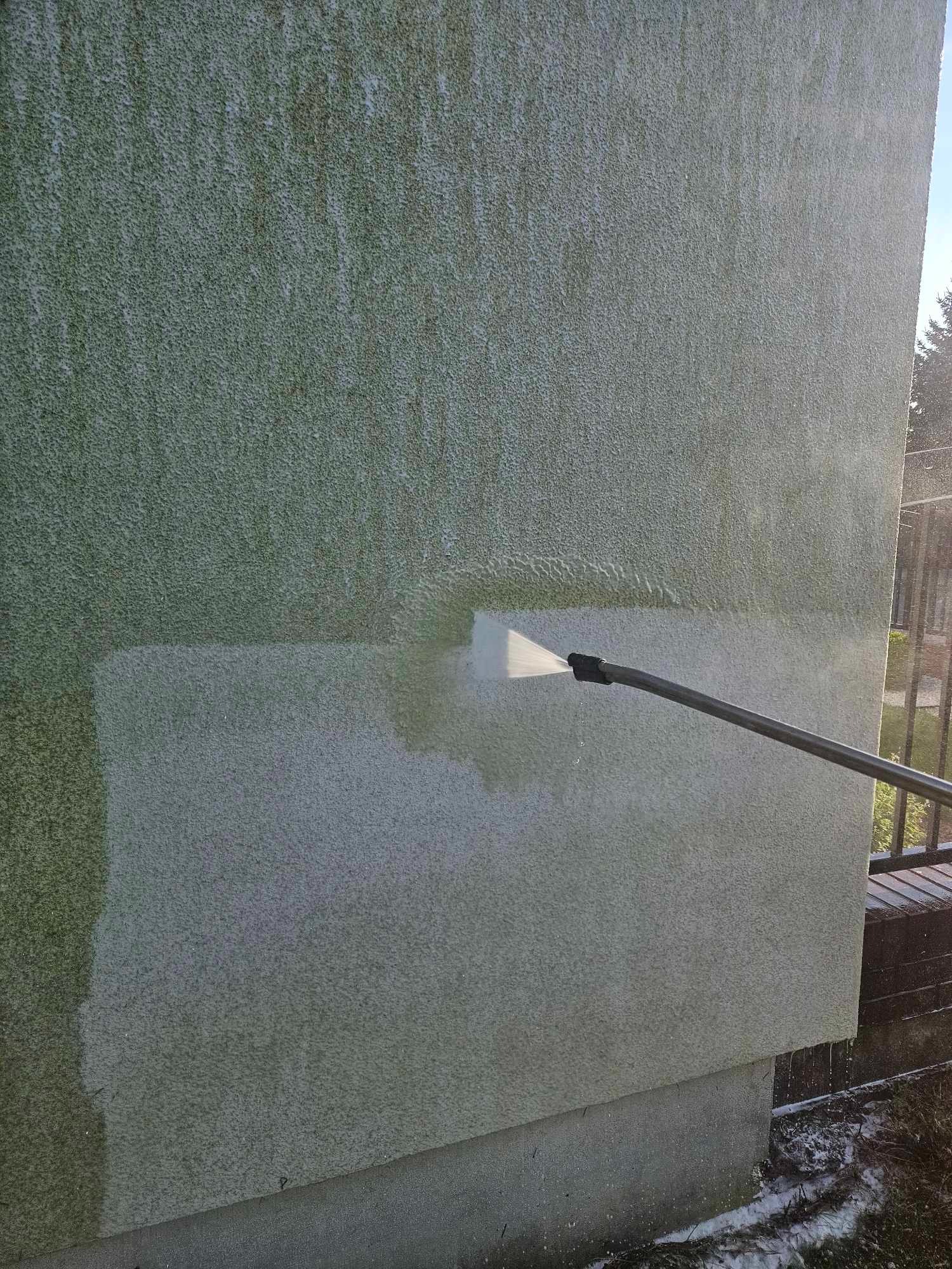 Strumień wody pod ciśnieniem z dyszy lancy czyści fragment zielonkawej, chropowatej elewacji budynku, widoczny kontrast czystej i zabrudzonej powierzchni.