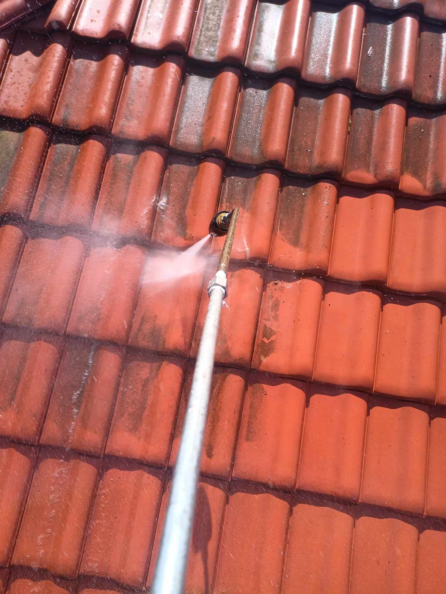 Czerwona dachówka ceramiczna poddawana czyszczeniu strumieniem wody pod ciśnieniem, widoczne różnice w kolorze między czystą a zabrudzoną powierzchnią.