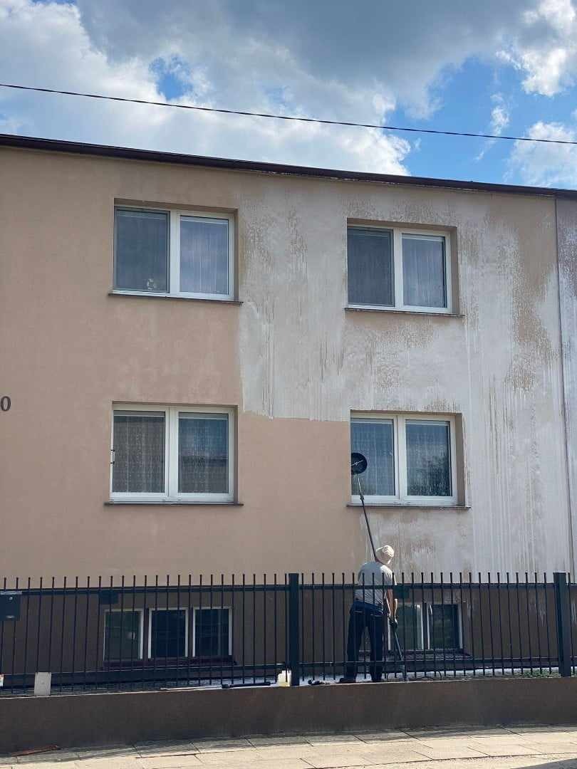 Pracownik z długim uchwytem czyści elewację budynku, widoczna różnica między umytą a brudną powierzchnią, cztery okna w kadrze.