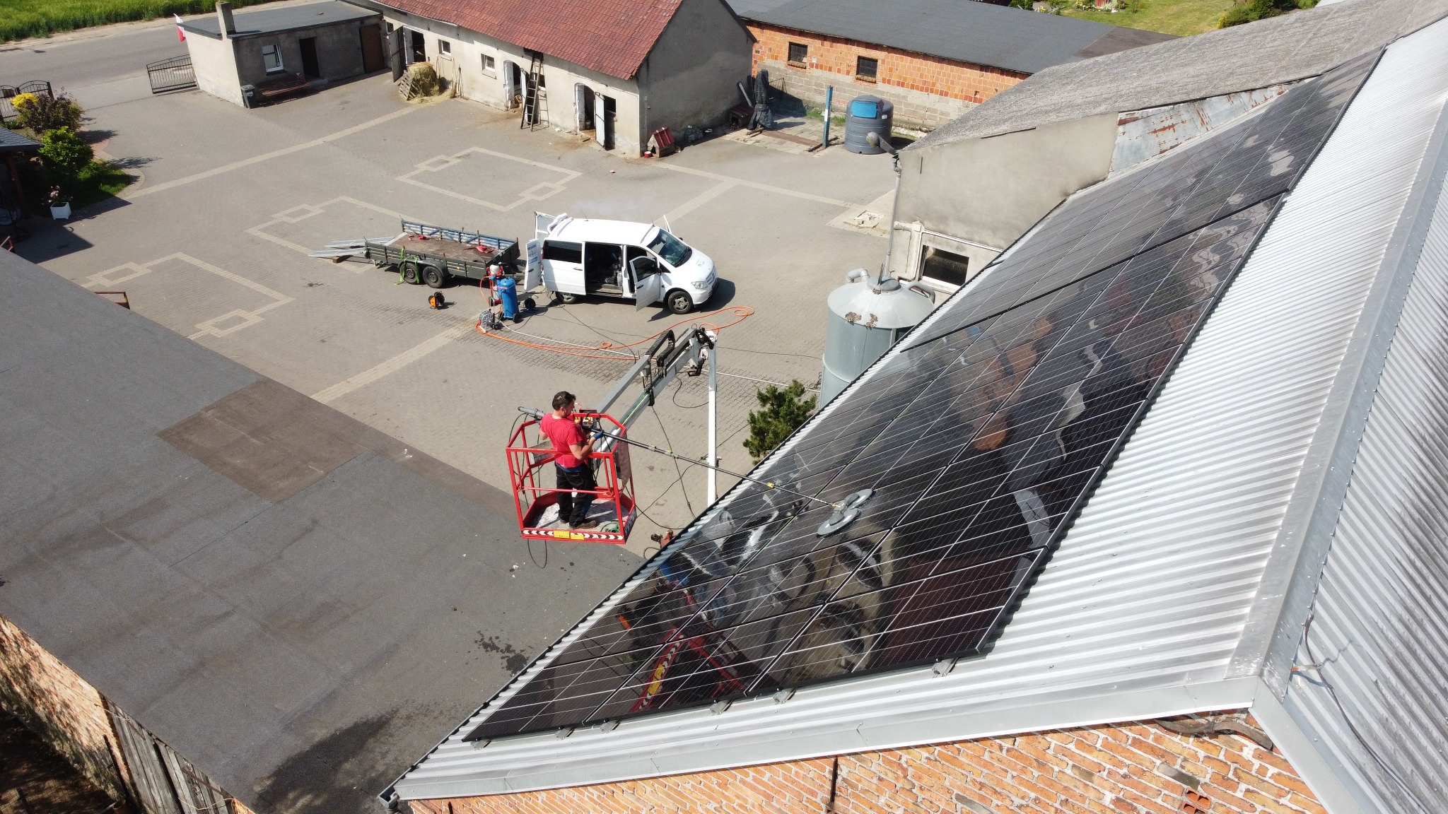 Pracownik na podnośniku koszowym czyści panele słoneczne na metalowym dachu budynku gospodarczego. Na dole widoczny biały samochód dostawczy i sprzęt do czyszczenia.