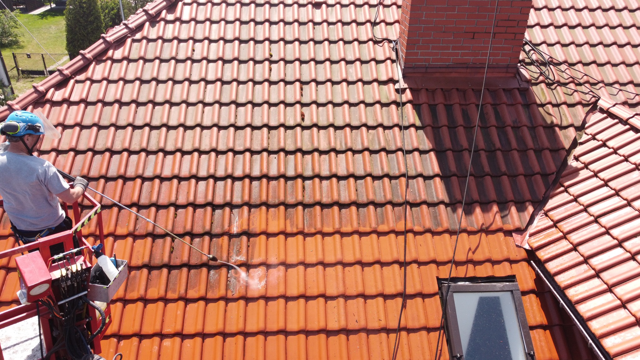 Częściowo wyczyszczony dach z czerwonej dachówki widziany z góry, pracownik na podnośniku koszowym czyści dach myjką ciśnieniową, widoczny komin i okno dachowe.