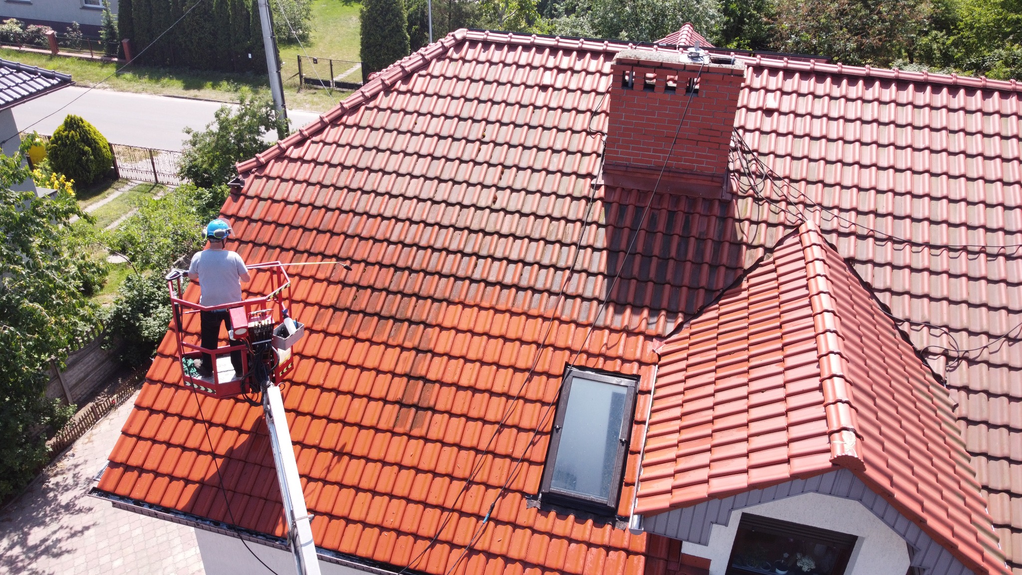 Pracownik na podnośniku koszowym czyści dach z czerwonej dachówki, widoczny fragment dachu przed i po czyszczeniu, ceglany komin.