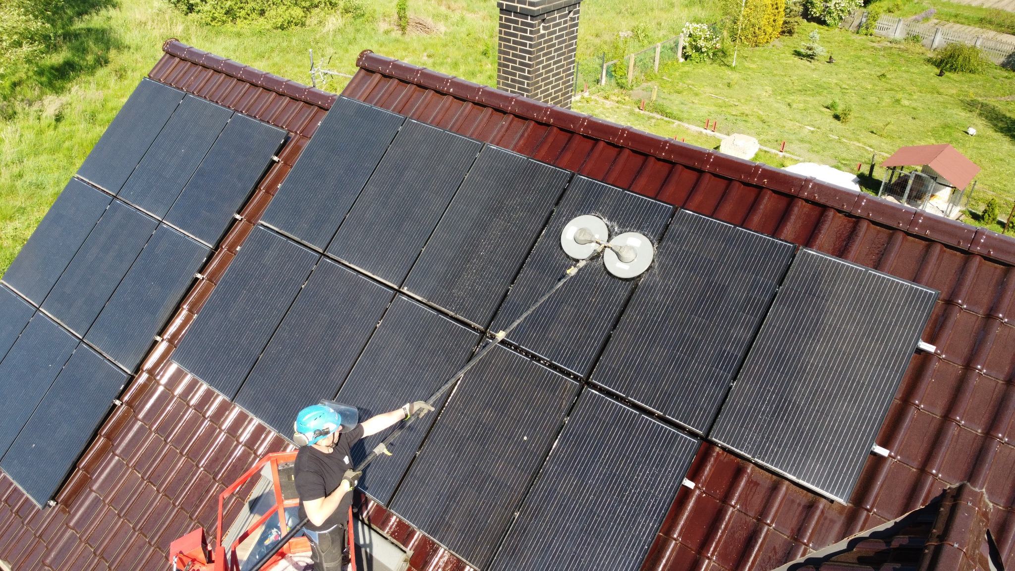 Pracownik w kasku i na podnośniku koszowym czyści panele słoneczne na brązowym dachu za pomocą specjalistycznej szczotki obrotowej, widok z góry.