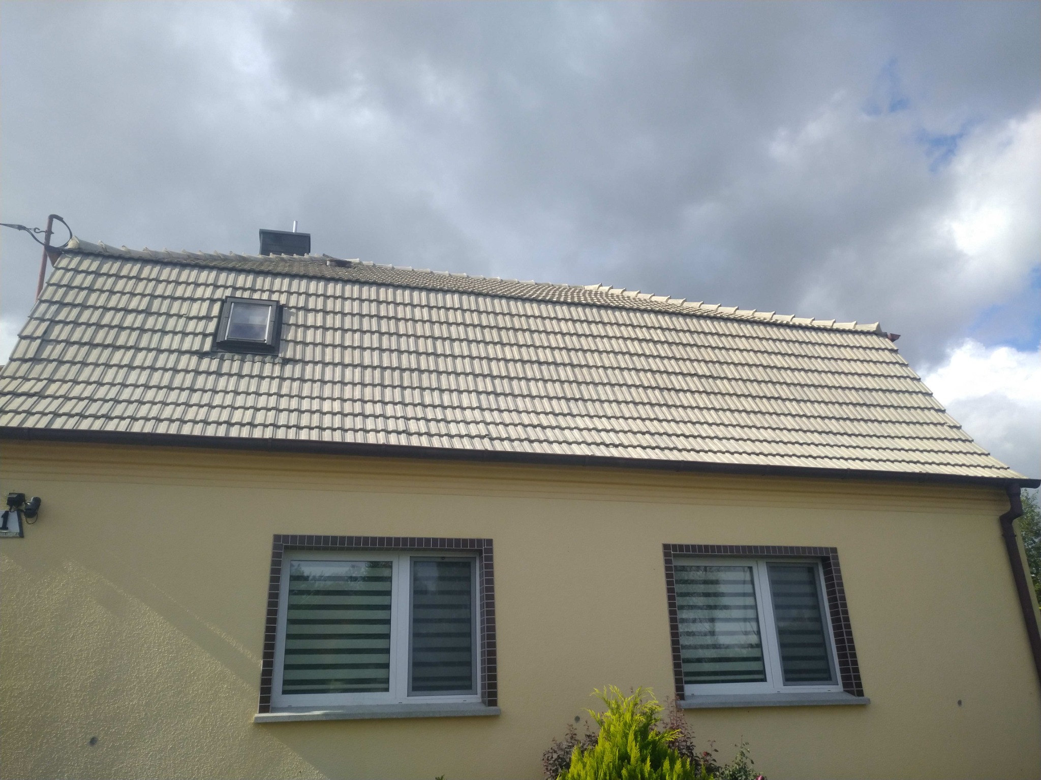 Dom jednorodzinny z beżową elewacją i dachem pokrytym jasną dachówką ceramiczną, widoczne okno dachowe i komin, pochmurne niebo w tle.