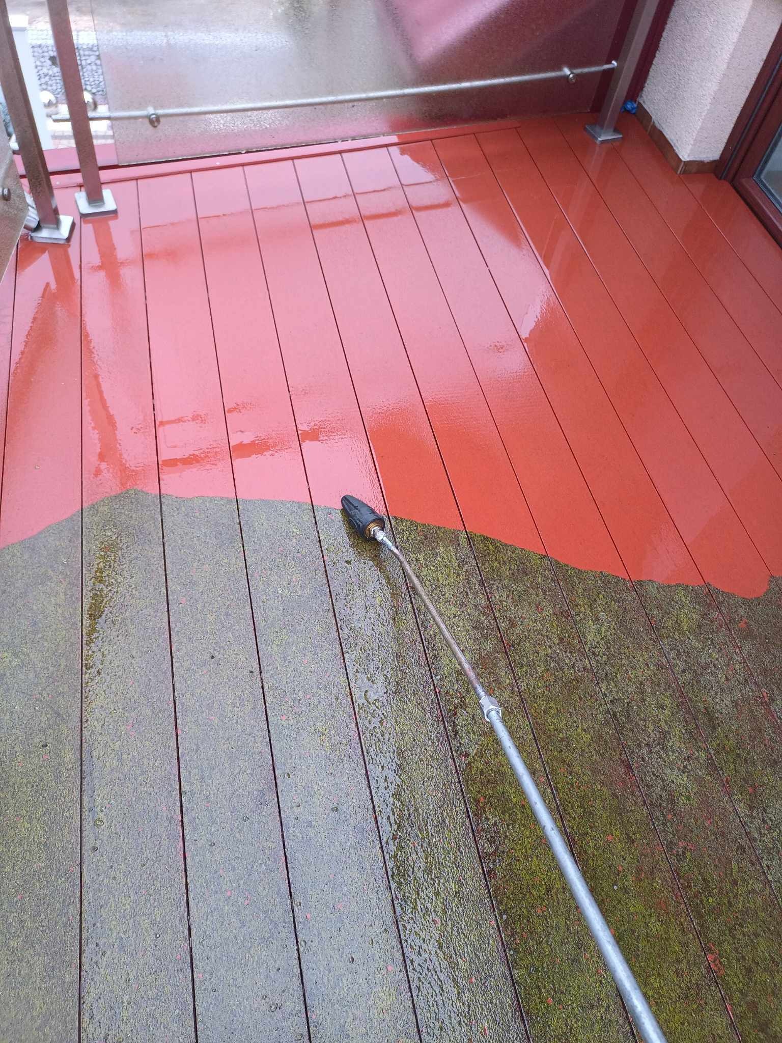 Częściowo wyczyszczony, czerwony drewniany taras z widoczną różnicą między zabrudzoną, zielonkawą częścią a czystą, czerwoną powierzchnią, czyszczony za pomocą myjki ciśnieniowej.