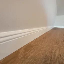 Nowoczesne wykończenie wnętrza: drewnopodobna podłoga, białe listwy przypodłogowe z frezowaniem, jasne ściany i gniazdko elektryczne w tle.