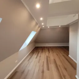 Poddasze po remoncie z drewnopodobną podłogą, skośnymi ścianami w odcieniach szarości, oknem dachowym i oświetleniem punktowym.