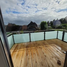 Firma Remontowo Budowlana Dominik Banaszak - Drewniany taras na balkonie z przeszkloną balustradą, widok na domy i pochmurne niebo, widoczne detale konstrukcyjne okna.