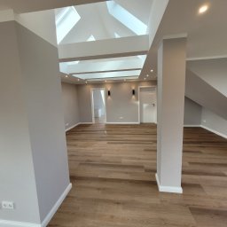 Firma Remontowo Budowlana Dominik Banaszak - Przestronne poddasze po remoncie z drewnopodobną podłogą, szarymi ścianami, białymi drzwiami i oknami dachowymi w suficie kasetonowym z oświetleniem punktowym.
