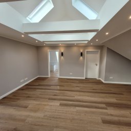 Firma Remontowo Budowlana Dominik Banaszak - Poddasze po remoncie z drewnopodobną podłogą, szarymi ścianami, białymi drzwiami i oknami dachowymi w suficie, oświetlone punktowymi lampami.