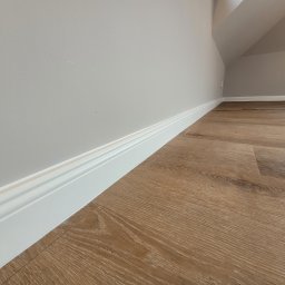 Firma Remontowo Budowlana Dominik Banaszak - Jasne panele podłogowe imitujące drewno, białe listwy przypodłogowe z frezowaniem, szara ściana pod skosem na poddaszu.