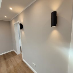 Firma Remontowo Budowlana Dominik Banaszak - Korytarz w Kotuni z drewnianą podłogą i szarą ścianą oświetloną przez dwa czarne, cylindryczne kinkiety.