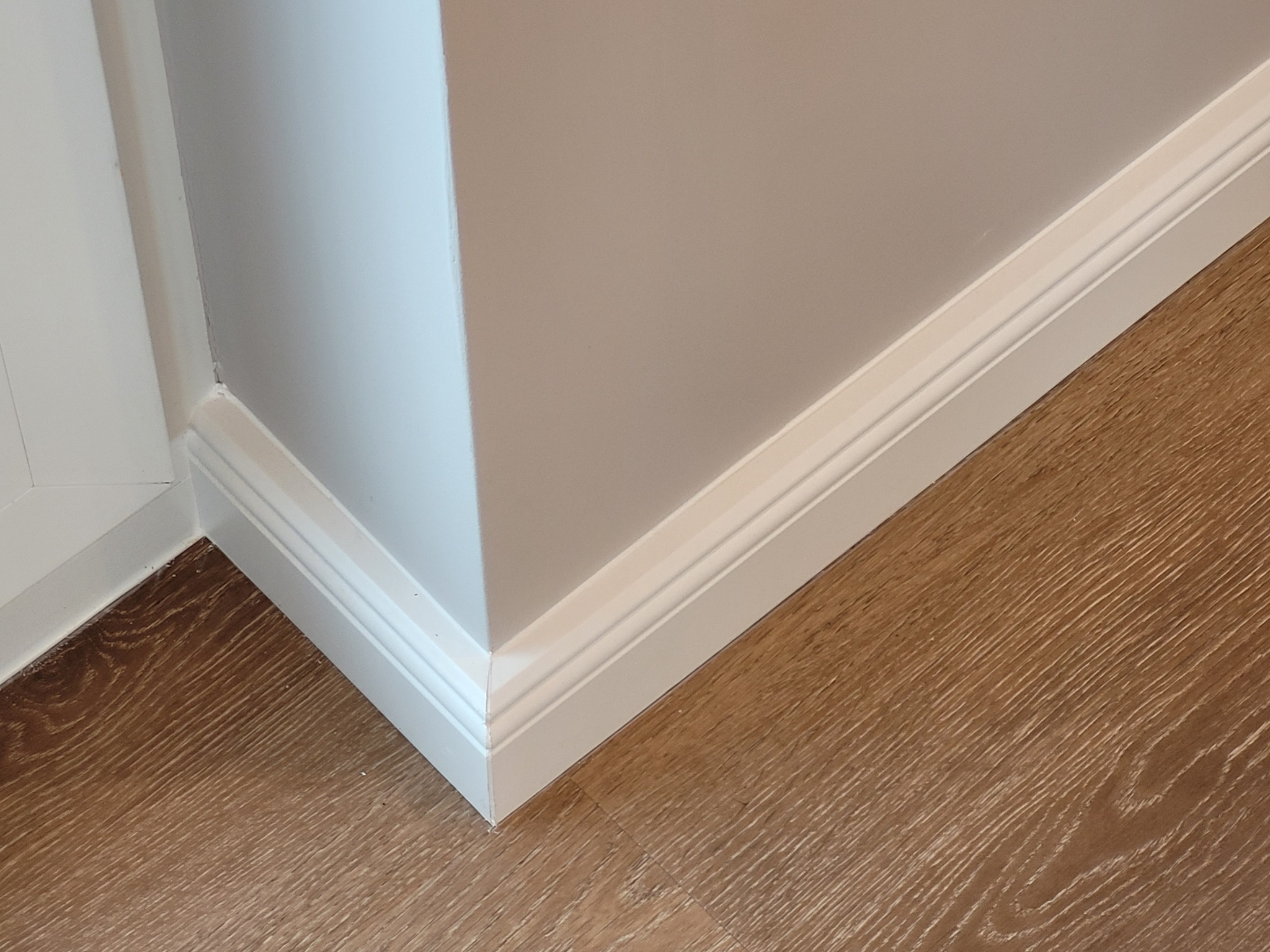 Detal wykończenia wnętrza: narożnik pomalowanej na szaro ściany, biała listwa przypodłogowa z frezowaniem i panel podłogowy imitujący jasne drewno.