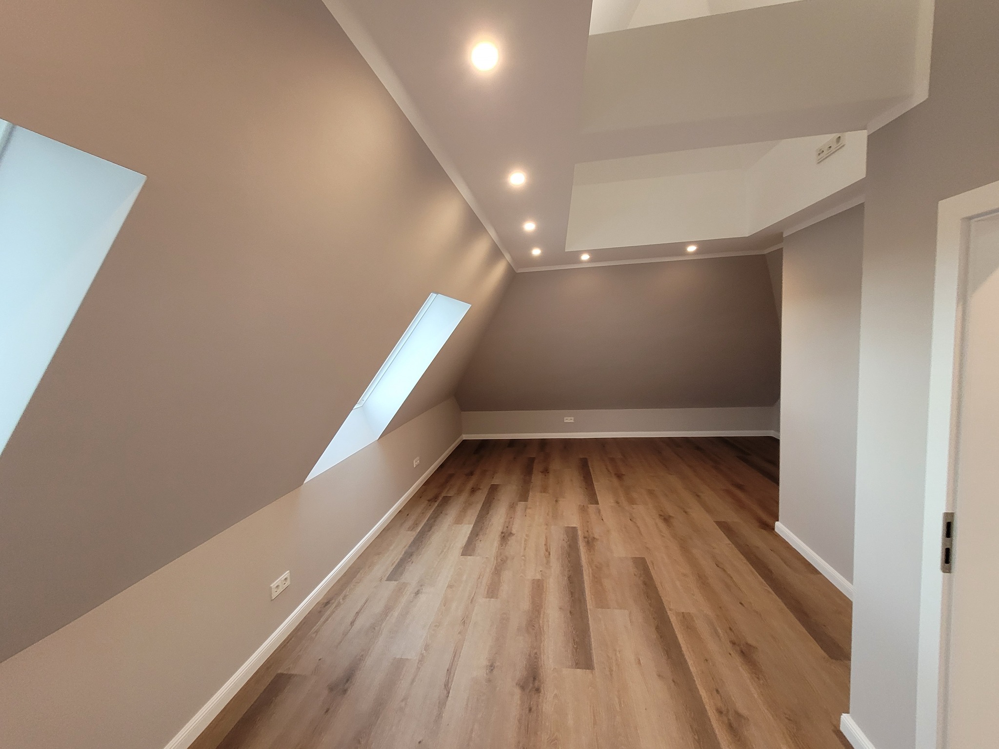 Poddasze po remoncie z drewnopodobną podłogą, skośnymi ścianami w odcieniach szarości, oknem dachowym i oświetleniem punktowym.